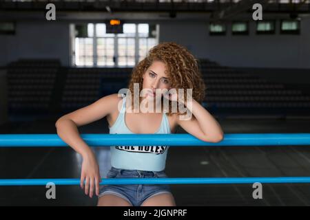 Charmante schlanke Frau mit lockigen Haaren, die auf die Kamera schaut, während sie sich an Metallstangen lehnt und Zeit im sportlichen Stadion verbringt Stockfoto