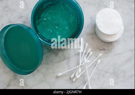 Feuchtigkeitsspendende Gelmaske im Kunststoffbehälter mit Baumwollpolstern und Stäbchen auf Marmortisch, Ansicht. Kosmetikprodukte zu Hause. Stockfoto