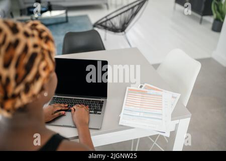 Nicht erkennbare afrikanische Frau, die von zu Hause aus arbeitet, von hinten geschossen und auf einen Laptop schaut. Laptop und Smartphone auf dem Schreibtisch des Heimbüros Stockfoto