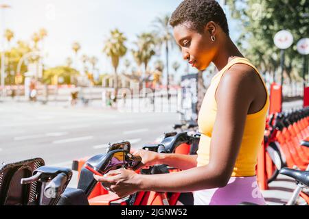 Trendige afroamerikanische Frau, die ihr Telefon benutzt, um das Fahrrad von einem Fahrradträger zu entsperren Stockfoto