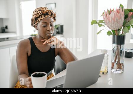 Junge Afrikanerin zu Hause, die ihre Online-Serie auf dem Laptop anschaut und einen Becher Kaffee hält Stockfoto