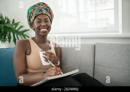 Afrikanerin studiert von zu Hause aus und schreibt Notizen in ein Buch. Entspannen auf dem Sofa zu Hause. Blick in die Kamera Stockfoto