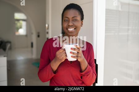 Porträt einer schönen afrikanischen Frau, die auf dem Weg zu Hause vor der Tür steht, einen Becher hält, lächelt und in die Kamera schaut Stockfoto