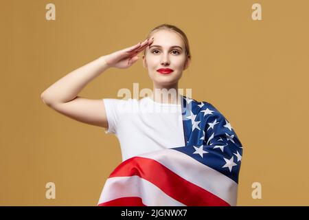 Attraktive Blondine posiert stolz mit einer amerikanischen Flagge. Fotoshooting im Studio auf gelbem Hintergrund. Stockfoto