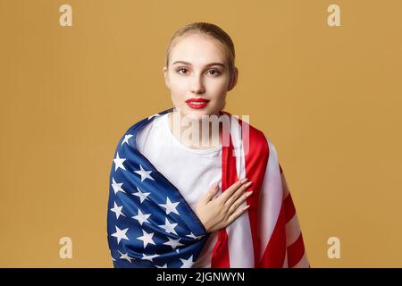 Attraktive Blondine posiert stolz mit einer amerikanischen Flagge. Fotoshooting im Studio auf gelbem Hintergrund. Stockfoto