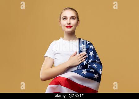 Attraktive Blondine posiert stolz mit einer amerikanischen Flagge. Fotoshooting im Studio auf gelbem Hintergrund. Stockfoto