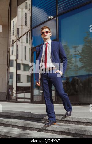Eleganter junger Mann in blauem Anzug, weißem Hemd, roter Krawatte, blauen Socken und Lederschuhen mit Holzsonnenbrille im modernen Firmengebäude Stockfoto