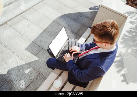 Junger Mann im Anzug mit Laptop mit leerem Bildschirm sitzt auf Steinbank. Blick von oben, Mockup. Mann mit Laptop für Online-Arbeit, Börse tr Stockfoto