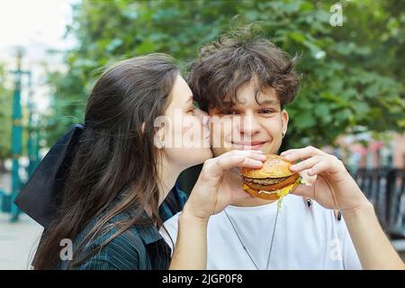 Ein junges Mädchen küsst einen Kerl, der einen Burger in den Händen hält. Stockfoto