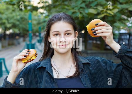 Die junge Brünette hält mit einem Lächeln zwei Hamburger in den Händen. Stockfoto