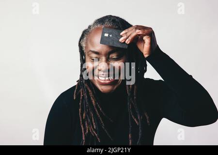 Eine fröhliche, reife Frau mit Dreadlocks lächelt fröhlich, während sie eine Kreditkarte vor ihre Stirn hält. Frau mittleren Alters, die elektronische Geräte empfiehlt Stockfoto