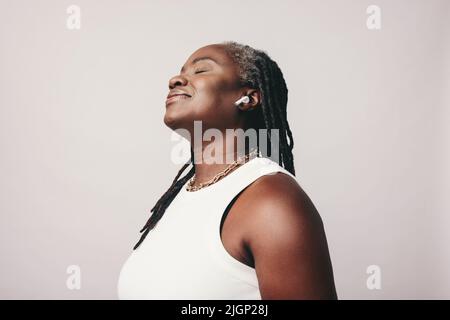 Sorglose Frau mit Dreadlocks, die Musik mit bluetooth-Ohrhörern hört. Reife Frau genießt ihre Lieblings-Wiedergabeliste, während sie gegen einen studi steht Stockfoto
