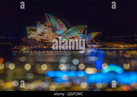 Doppelbelichtungsfoto des Opernhauses von Sydney, einem australischen Wahrzeichen, das während des Vivid Festival 2022 beleuchtet wurde, mit Bokeh-Effekt im Vordergrund. Stockfoto