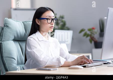 Junge asiatische Studentin in Brille sitzt und studiert online am Computer zu Hause am Tisch, tippt auf der Tastatur, besteht die Prüfung, ernst. Stockfoto