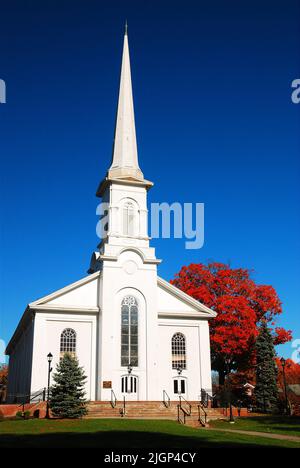 Eine klassische Kirche im New England Stil mit einem hohen Kirchturm steht hoch in einer kleinen Stadt, umgeben von Herbstblättern und saisonaler Herbstfarbe Stockfoto