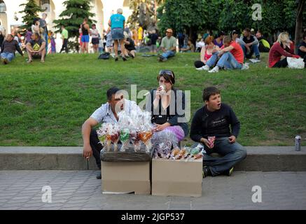 Mann, Frau und Junge Straßenhändler verkaufen Süßigkeiten sitzen auf dem Bürgersteig, verschwommene Menschen entspannen sich auf einem Gras im Stadtpark. 19.Mai 2019. Kiew, Ukr Stockfoto