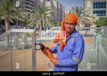 Porträt eines jungen Straßenarbeiters aus dem Nahen Osten mit blauer Uniform und orangefarbenem Kopfschmuck und polierfähigem Polierwerkzeug in den Händen.VAE Stockfoto