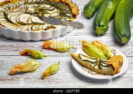 Offene Kuchenform aus Zucchini und Frischkäse auf einem weißen Teller und in einer von Blumen umgebenen Backform auf einem hellen, bemalten Holztisch. Vorderansicht Stockfoto