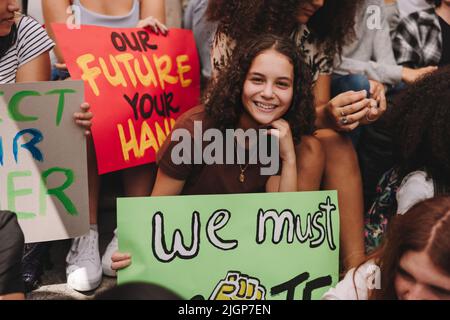 Ein fröhliches Teenager-Mädchen lächelt an der Kamera und hält ein Banner, während es mit einer Gruppe von Klimaaktivisten sitzt. Multikulturelle Demonstranten, die für c streiken Stockfoto