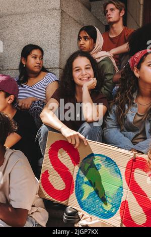 Ein glückliches Teenager-Mädchen mit einem Transparent „Save our species“, während es auf einer Demonstration zum Klimawandel sitzt. Eine Gruppe multikultureller Jugendaktivisten protestiert Stockfoto
