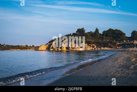 Blick auf den Küstenstrand im Hintergrund ein Felsvorsprung, der mit grünen Bäumen und Sträuchern bedeckt ist. Ormos Panagias, Griechenland. Stockfoto