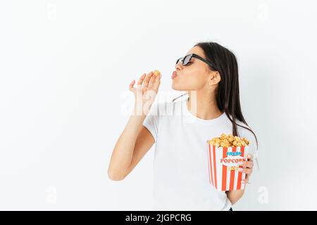 Junge Frau, die gerne Popcorn isst, steht mit 3D Gläsern auf weißem Studiohintergrund Stockfoto