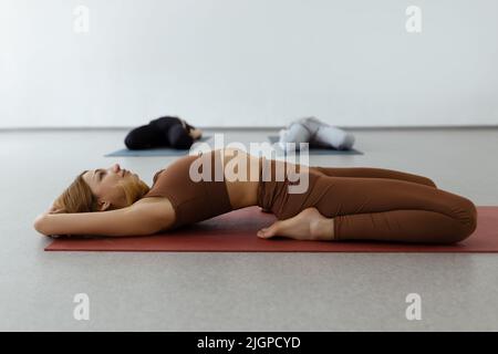 Schlanke junge Frau, die in Supta Virasana liegt, in der Liegenden Heldenpose, die Bauch- und Rückenmuskulatur im Fitnessstudio auf der Matte dehnt Stockfoto