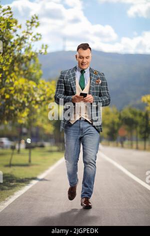 Ganzkörperporträt eines Mannes in stilvollem Anzug und Jeans, der auf einer Fußgängerstraße unterwegs ist und ein Smartphone verwendet Stockfoto