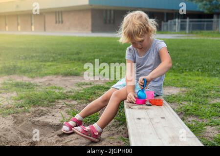 Kleines Kind, das am sonnigen Tag draußen auf dem Schulhof mit Sand und Spielzeug spielt. Kleines Mädchen, das im Sommer auf der Bank auf dem Spielplatz sitzt Stockfoto