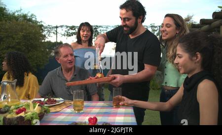 Der Grillkoch bringt Essen zu Freunden und Familie, die sich um den Tisch versammelt haben. BBQ-Koch serviert Essen an den Vater Stockfoto