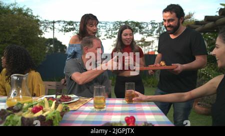 Der Grillkoch bringt Essen zu Freunden und Familie, die sich um den Tisch versammelt haben. BBQ-Koch serviert Essen an den Vater Stockfoto