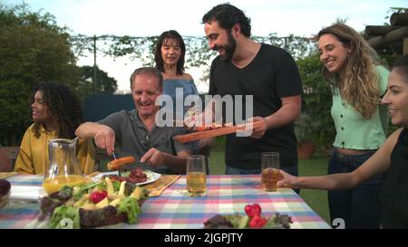 Der Grillkoch bringt Essen zu Freunden und Familie, die sich um den Tisch versammelt haben. BBQ-Koch serviert Essen an den Vater Stockfoto