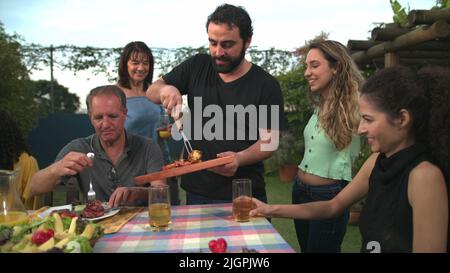 Der Grillkoch bringt Essen zu Freunden und Familie, die sich um den Tisch versammelt haben. BBQ-Koch serviert Essen an den Vater Stockfoto