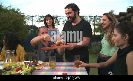 Der Grillkoch bringt Essen zu Freunden und Familie, die sich um den Tisch versammelt haben. BBQ-Koch serviert Essen an den Vater Stockfoto