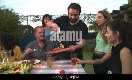 Der Grillkoch bringt Essen zu Freunden und Familie, die sich um den Tisch versammelt haben. BBQ-Koch serviert Essen an den Vater Stockfoto