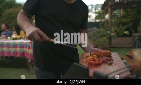 Der Grillkoch bringt Essen zu Freunden und Familie, die sich um den Tisch versammelt haben. BBQ-Koch serviert Essen an den Vater Stockfoto