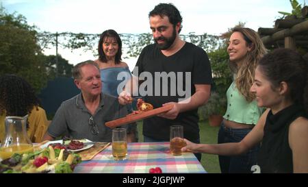 Der Grillkoch bringt Essen zu Freunden und Familie, die sich um den Tisch versammelt haben. BBQ-Koch serviert Essen an den Vater Stockfoto