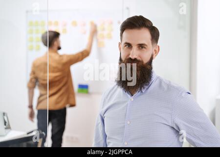 Glückliches Lächeln bärtige Senior Developer oder Manager in modernen IT-Büro. Ein weiterer Profi, der mit Scrum Desk im Hintergrund arbeitet. Erfolgreich Stockfoto