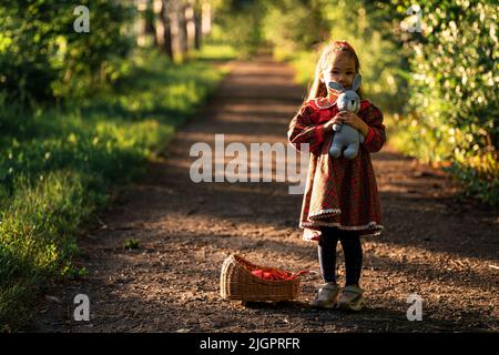 Kleines Mädchen in rotem Kleid hält Puppe neben Weidenwiege Stockfoto