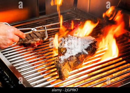 Gegrilltes Rinderschnitzel mit Salz und Flammen des Feuers und Hand mit Zange Stockfoto
