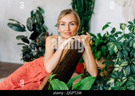 Porträt einer jungen lächelnden Frau, die auf einem Holzstuhl zwischen Pflanzen sitzt. Stockfoto