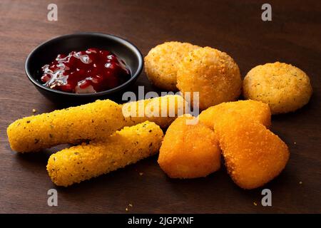 Gebratener panierter Brie- und Camembert-Nuggets, Mozzarella-Sticks und Preiselbeersauce auf dunklem Holz. Stockfoto