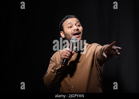Emotionaler indischer Komiker mit offenem Mund, das Mikrofon hält und während des Monologes mit dem Finger auf Schwarz zeigt Stockfoto