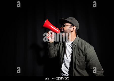 Seitenansicht eines emotionalen Hip-Hop-Darstellers aus dem Nahen Osten mit Sonnenbrille und Kappe, die Lautsprecher auf Schwarz singen und halten Stockfoto