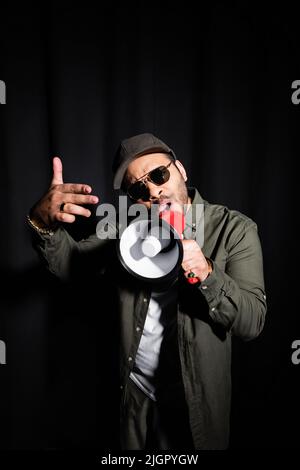 Emotionaler Hip-Hop-Performer aus dem Nahen Osten in Sonnenbrillen und Mütze, der in einem Megaphon auf Schwarz spricht Stockfoto