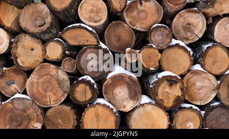 Gestapelte, mit Schnee bedeckte Holzstämme, die in mehreren Reihen ausgelegt sind. Natürlicher Hintergrund aus Holz. Nahaufnahme des Brennholzes. Stockfoto