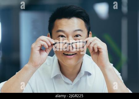 Überrascht asiatische Mann schießt Brille schockiert Blick auf Kamera. Fröhliches Lächeln freut sich. Schauen Sie sich Monitor-Bildschirm Webcam-Ansicht. Erstaunt Nahaufnahme Porträt. Gesicht hübscher Mann Büroangestellter Wow Ausdruck Stockfoto