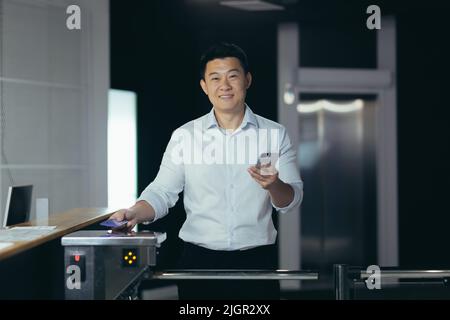 Porträt eines asiatischen Geschäftsmannes, der zum ersten Mal zur Arbeit kommt, ins Büro geht, lächelt und blickt auf die Kamera an der Eingangstür, hält ein Telefon in den Händen Stockfoto