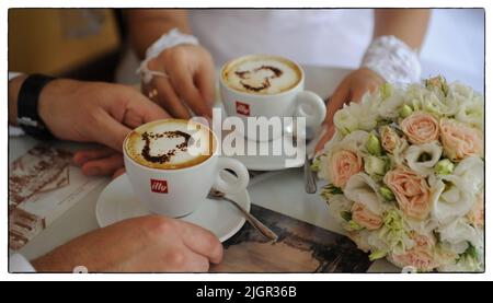 Hochzeitshände eines Mannes und einer Frau der Braut und des Bräutigams in den Händen halten sie zwei Tassen Kaffee, auf denen sich Schaum und ein gezogenes Herz auf einem Tisch mit weißer Tischdecke befinden. Hochwertige Fotos Stockfoto