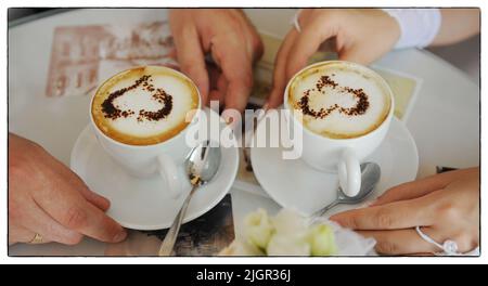 Hochzeitshände eines Mannes und einer Frau der Braut und des Bräutigams in den Händen halten sie zwei Tassen Kaffee, auf denen sich Schaum und ein gezogenes Herz auf einem Tisch mit weißer Tischdecke befinden. Hochwertige Fotos Stockfoto